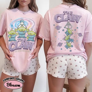 May include: A light pink t-shirt featuring a graphic of three green aliens and a claw machine mechanism. The front displays the text THE CLAW and THE MYSTIC PORTAL AWAITS. The back shows the aliens hanging from a claw with planets and stars.