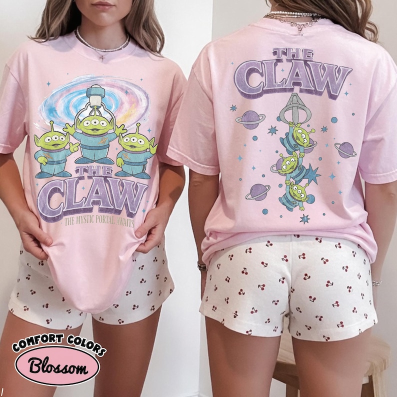 May include: A light pink t-shirt featuring a graphic of three green aliens and a claw machine mechanism. The front displays the text THE CLAW and THE MYSTIC PORTAL AWAITS. The back shows the aliens hanging from a claw with planets and stars.