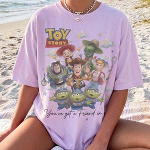 Puede incluir: Una camiseta de manga corta de color morado claro con un estampado gr&aacute;fico de los personajes de Toy Story, incluyendo a Woody, Buzz Lightyear, Jessie, Rex, Bo Peep y los alien&iacute;genas verdes. El texto "TOY STORY" y "You've got a Friend in" aparece.