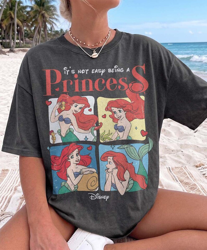 Puede incluir: Una camiseta de manga corta gris carb&oacute;n con un estampado gr&aacute;fico del personaje Ariel de La Sirenita en cuatro paneles distintos. El texto "It's not easy being a Princess" est&aacute; impreso en rojo y blanco sobre el logotipo de Disney.
