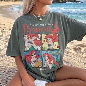 Puede incluir: Una camiseta de manga corta color verde salvia con un estampado gr&aacute;fico de Ariel de La Sirenita en cuatro paneles distintos. El texto dice "It's not easy being a Princess" en letras rojas y blancas, con el logotipo de Disney en la parte inferior.