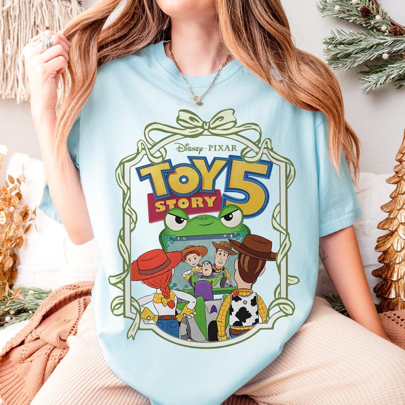 May include: Light blue t-shirt featuring a Disney Pixar Toy Story 5 graphic. The design includes the movie title, a green frog, and several characters. The shirt is a casual wear item.
