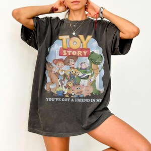 K&ouml;nnte beinhalten: Dunkelgraues T-Shirt mit dem Toy Story-Filmlogo und -Charakteren. Die Grafik zeigt Woody, Buzz Lightyear und andere Charaktere mit dem Text "YOU'VE GOT A FRIEND IN ME".