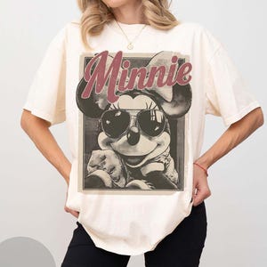 May include: A cream-coloured short-sleeve t-shirt featuring a graphic print of Minnie Mouse wearing dark sunglasses. The word Minnie is printed in a large, distressed red font above the character. The design is enclosed in a rectangular frame.