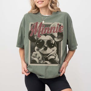 May include: An olive green short-sleeve t-shirt featuring a graphic print of Minnie Mouse wearing sunglasses. The word Minnie is printed in a large, dark red script font above the character. The design is enclosed within a rectangular border.