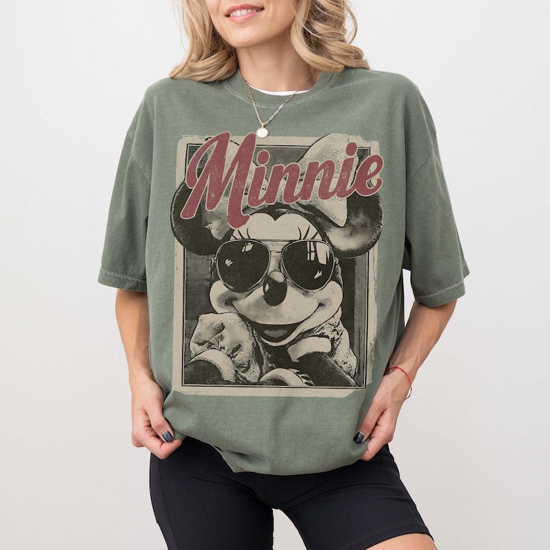 May include: An olive green short-sleeve t-shirt featuring a graphic print of Minnie Mouse wearing sunglasses. The word Minnie is printed in a large, dark red script font above the character. The design is enclosed within a rectangular border.
