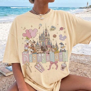 May include: A pale yellow short-sleeve t-shirt featuring a graphic print of a castle, Mickey Mouse, Minnie Mouse, Goofy, Donald Duck, and Daisy Duck. The design includes the text Disneyland, balloons, popcorn, a bow, and various small icons.