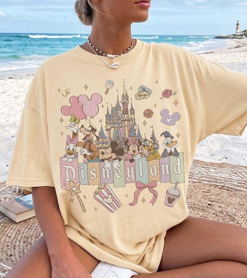May include: A pale yellow short-sleeve t-shirt featuring a graphic print of a castle, Mickey Mouse, Minnie Mouse, Goofy, Donald Duck, and Daisy Duck. The design includes the text Disneyland, balloons, popcorn, a bow, and various small icons.