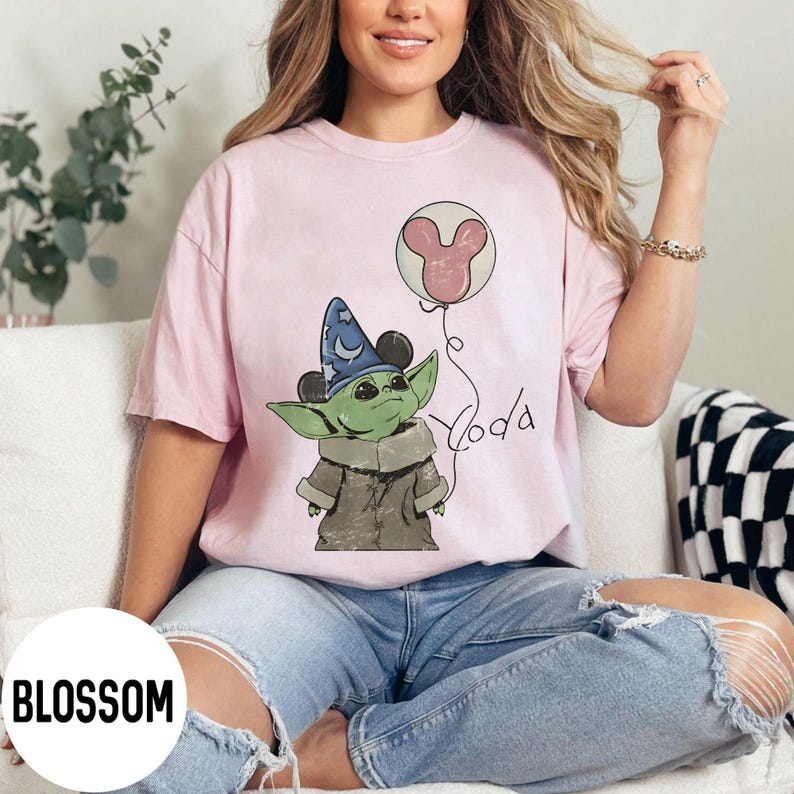 K&ouml;nnte beinhalten: Hellrosa T-Shirt mit einer Cartoon-Illustration von Grogu, der einen Zaubererhut mit Mickey Mouse-Ohren tr&auml;gt und einen Mickey Mouse-Ballon h&auml;lt. Das Wort "Yoda" ist in einer Schriftart geschrieben. Das T-Shirt hat eine lockere Passform.