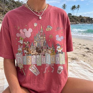 May include: A dusty rose short-sleeve t-shirt featuring a graphic print of a castle, Mickey Mouse, Minnie Mouse, Donald Duck, Daisy Duck, Goofy, and Pluto. The text "Disneyland" is printed in a serif font across the centre with various icons.
