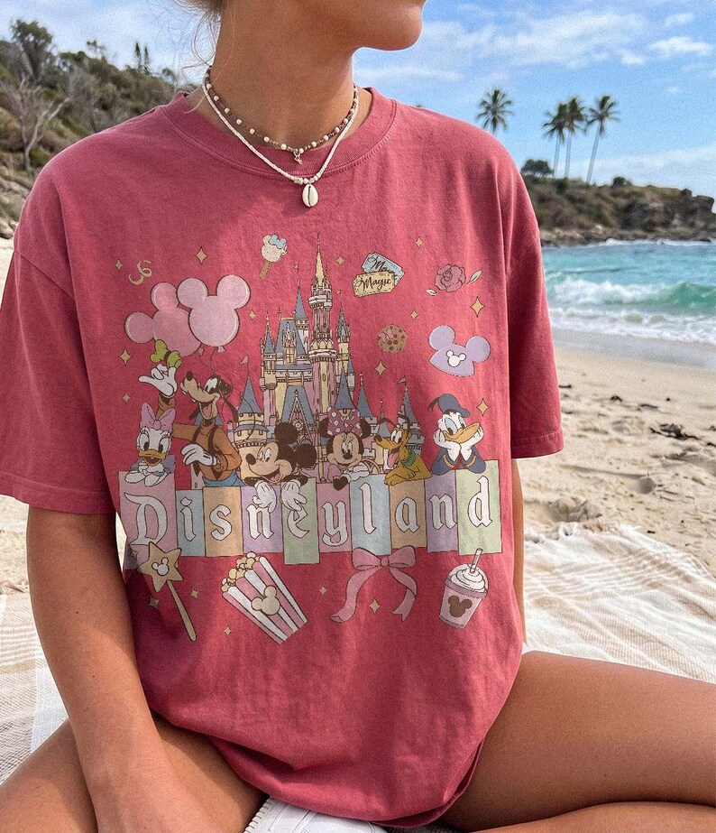May include: A dusty rose short-sleeve t-shirt featuring a graphic print of a castle, Mickey Mouse, Minnie Mouse, Donald Duck, Daisy Duck, Goofy, and Pluto. The text "Disneyland" is printed in a serif font across the centre with various icons.