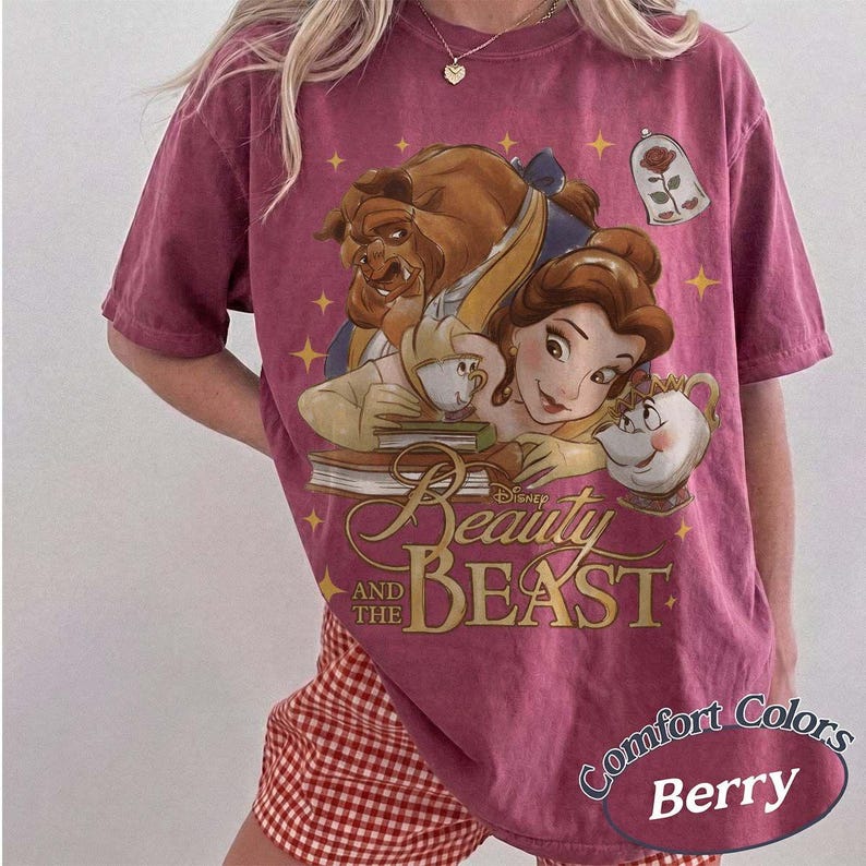 May include: A berry-coloured short-sleeve t-shirt featuring a graphic print of Belle, the Beast, Mrs. Potts, and Chip from Beauty and the Beast. The design includes a rose in a glass dome, yellow stars, and the text Disney Beauty and the Beast.