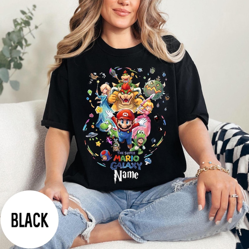 Puede incluir: Camiseta negra con un gr&aacute;fico colorido de personajes de Super Mario Galaxy, incluyendo a Mario, la Princesa Peach y Bowser. El dise&ntilde;o se destaca sobre un fondo negro. El texto "THE SUPER MARIO GALAXY Name" tambi&eacute;n es visible.