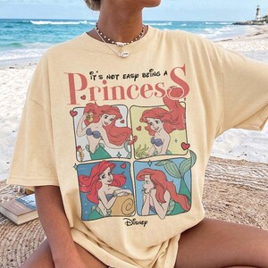 Puede incluir: Una camiseta de manga corta de color amarillo p&aacute;lido con un estampado gr&aacute;fico del personaje Ariel de La Sirenita en cuatro paneles distintos. El texto sobre el gr&aacute;fico dice, It's not easy being a Princess, con el logotipo de Disney abajo.