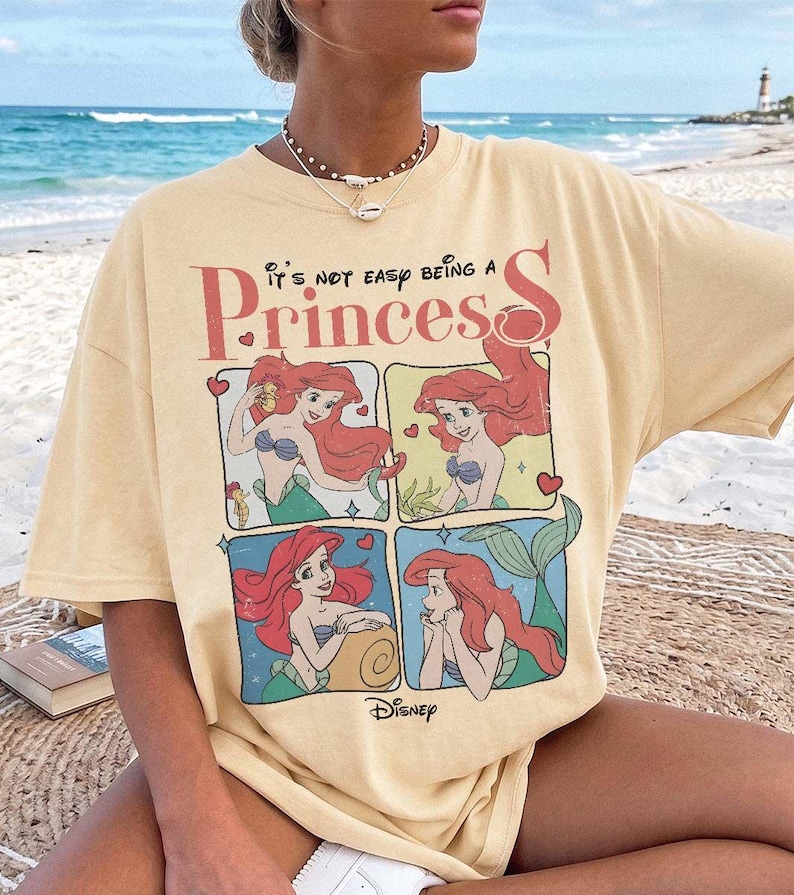 Puede incluir: Una camiseta de manga corta de color amarillo p&aacute;lido con un estampado gr&aacute;fico del personaje Ariel de La Sirenita en cuatro paneles distintos. El texto sobre el gr&aacute;fico dice, It's not easy being a Princess, con el logotipo de Disney abajo.
