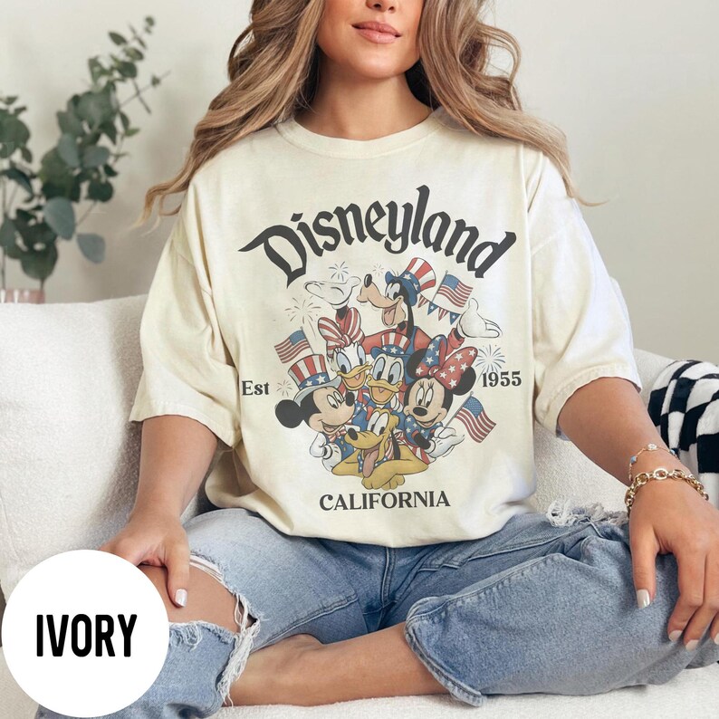May include: Ivory-colored t-shirt featuring a graphic of Disney characters, including Mickey Mouse, Minnie Mouse, Donald Duck, and Goofy, with American flags. The shirt has the text "Disneyland California Est 1955".