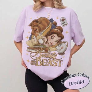 May include: A light purple t-shirt featuring a graphic print of Belle and the Beast from Beauty and the Beast, along with Mrs. Potts, Chip, and a rose in a glass dome. The text Beauty and the Beast is printed in gold below the characters.