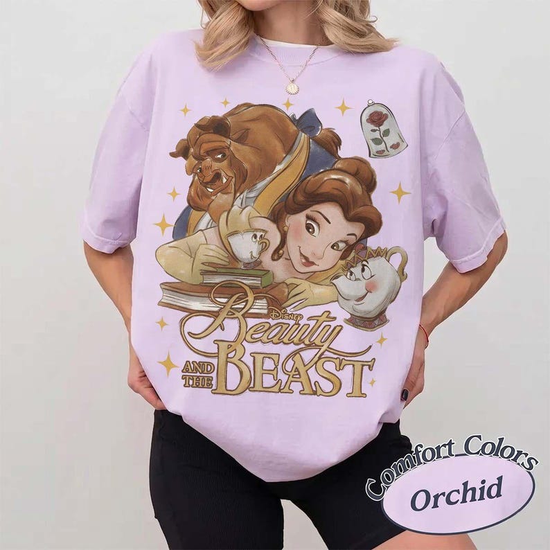 May include: A light purple t-shirt featuring a graphic print of Belle and the Beast from Beauty and the Beast, along with Mrs. Potts, Chip, and a rose in a glass dome. The text Beauty and the Beast is printed in gold below the characters.