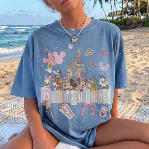 May include: A blue short-sleeve t-shirt featuring a graphic print of Disneyland with Mickey Mouse, Minnie Mouse, Goofy, Donald Duck, and Daisy Duck. The design includes a castle, balloons, popcorn, and the text Disneyland in a pastel colour palette.