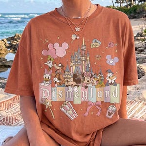 May include: A rust-coloured short-sleeve t-shirt featuring a graphic print of the Disneyland castle, Mickey Mouse, Minnie Mouse, Goofy, Donald Duck, and Daisy Duck. The design includes pastel-coloured blocks, balloons, popcorn, and a bow.