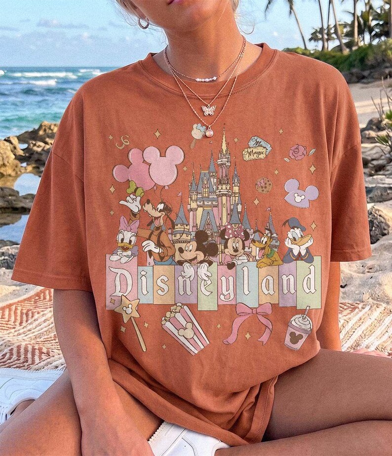 May include: A rust-coloured short-sleeve t-shirt featuring a graphic print of the Disneyland castle, Mickey Mouse, Minnie Mouse, Goofy, Donald Duck, and Daisy Duck. The design includes pastel-coloured blocks, balloons, popcorn, and a bow.