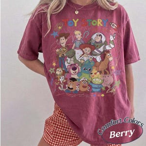 May include: A berry-colored short-sleeve t-shirt featuring a graphic print of Toy Story characters including Woody, Buzz Lightyear, Jessie, and others. The design includes the text TOY STORY in colorful letters and various hand-drawn style doodles.