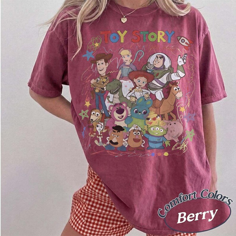 May include: A berry-colored short-sleeve t-shirt featuring a graphic print of Toy Story characters including Woody, Buzz Lightyear, Jessie, and others. The design includes the text TOY STORY in colorful letters and various hand-drawn style doodles.
