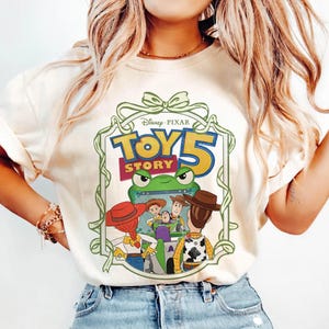 May include: Cream-colored t-shirt featuring the Disney Pixar Toy Story 5 logo in a green and yellow frame. The graphic includes images of Woody, Buzz Lightyear, and other characters from the movie. The shirt has short sleeves and a crew neck.