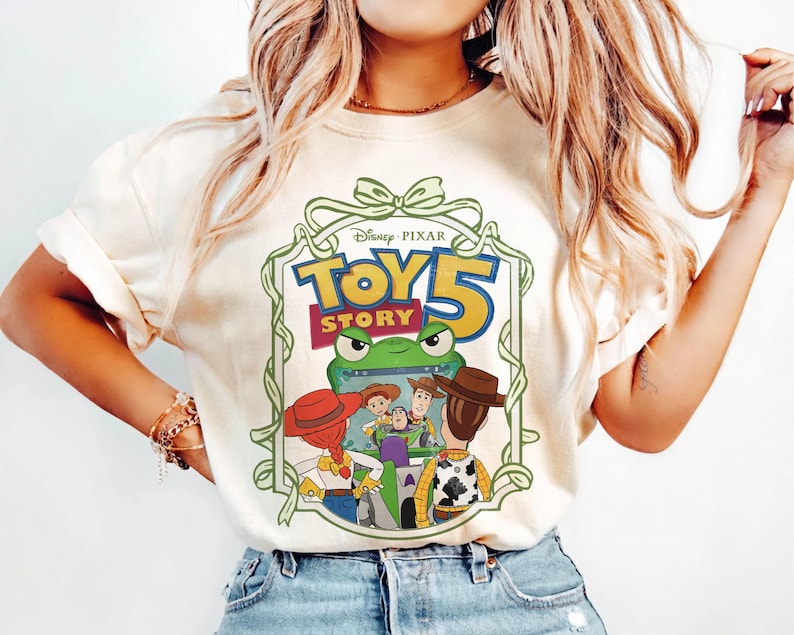 May include: Cream-colored t-shirt featuring the Disney Pixar Toy Story 5 logo in a green and yellow frame. The graphic includes images of Woody, Buzz Lightyear, and other characters from the movie. The shirt has short sleeves and a crew neck.