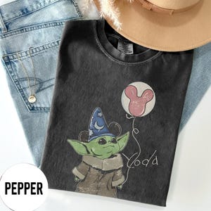 K&ouml;nnte beinhalten: Dunkelgraues T-Shirt mit einer Cartoonfigur mit blauem Zaubererhut und rosa Mickey Mouse-Ballon. Das Wort "Yoda" ist in einer verspielten Schrift geschrieben. Das Shirt liegt auf einer blauen Jeans und einem beigen Hut.