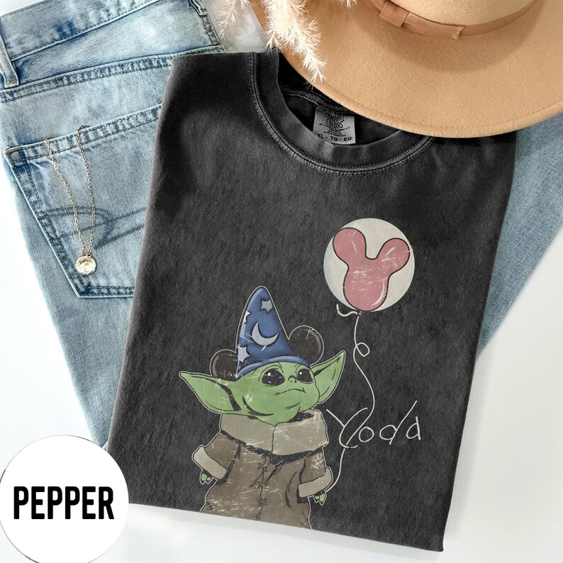 K&ouml;nnte beinhalten: Dunkelgraues T-Shirt mit einer Cartoonfigur mit blauem Zaubererhut und rosa Mickey Mouse-Ballon. Das Wort "Yoda" ist in einer verspielten Schrift geschrieben. Das Shirt liegt auf einer blauen Jeans und einem beigen Hut.