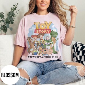 K&ouml;nnte beinhalten: Hellrosa T-Shirt mit dem Schriftzug "TOY STORY" und einer Grafik von Figuren aus dem Film. Darunter steht der Text "YOU'VE GOT A FRIEND IN ME". Das T-Shirt wird getragen.