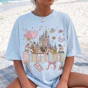 May include: A light blue short-sleeve t-shirt featuring a graphic print of a castle, Mickey Mouse, Minnie Mouse, Donald Duck, Daisy Duck, Goofy, and Pluto. The text Disneyland is printed in a stylised font above icons of popcorn and a bow.
