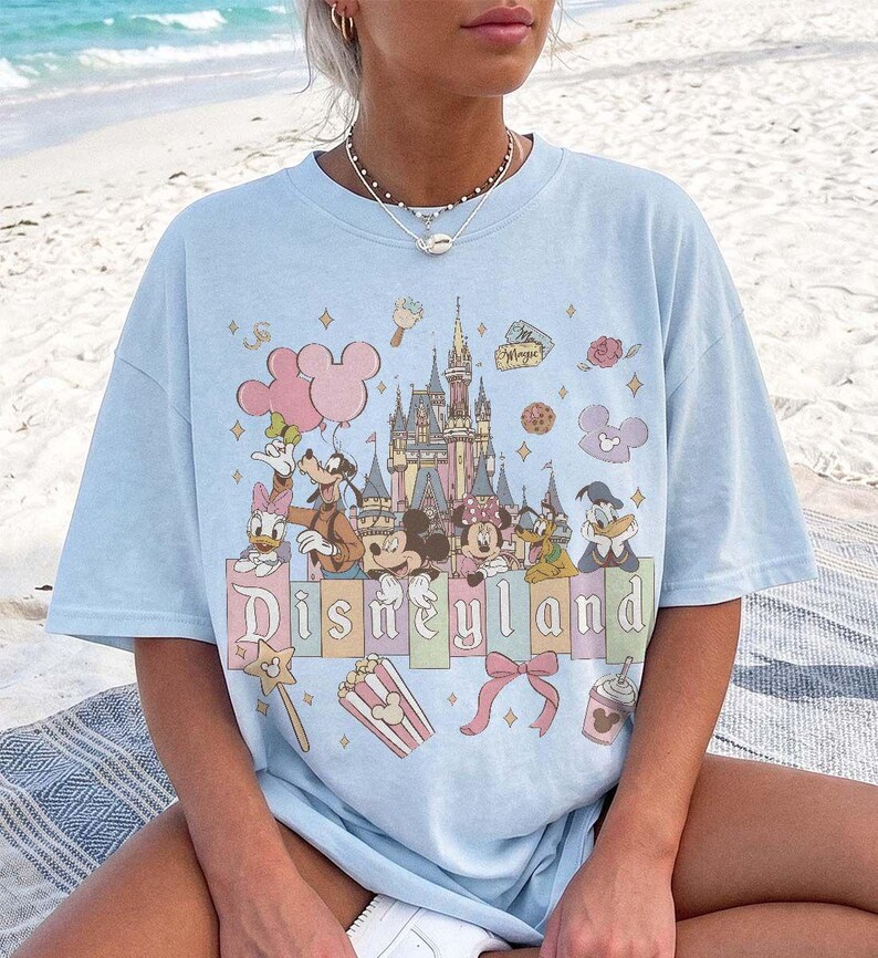 May include: A light blue short-sleeve t-shirt featuring a graphic print of a castle, Mickey Mouse, Minnie Mouse, Donald Duck, Daisy Duck, Goofy, and Pluto. The text Disneyland is printed in a stylised font above icons of popcorn and a bow.