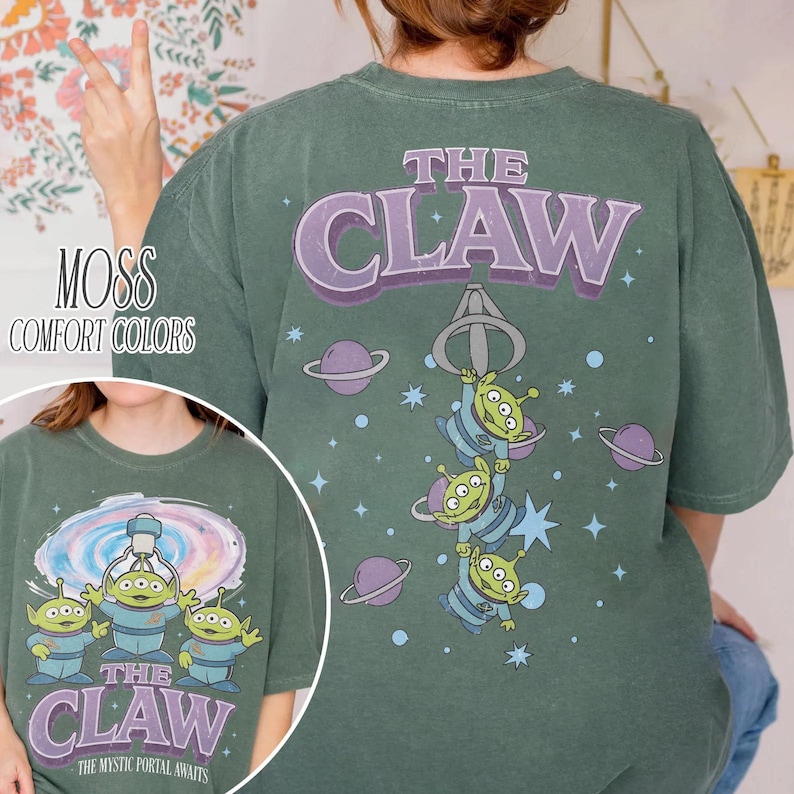 May include: A moss green t-shirt featuring a graphic of three green three-eyed aliens and a claw machine mechanism. The back design includes the text THE CLAW with purple lettering, planets, and stars. The front design shows the same aliens.