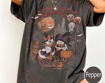 Könnte beinhalten: Ein anthrazitfarbenes Kurzarm-T-Shirt mit einem Grafikdruck von zwei Zeichentrickfiguren in Hexenhüten und gestreiften Socken. Das Design zeigt einen großen orangefarbenen Mond, ein Schloss, Kürbisse, Spinnennetze und eine rote Schleife oben.