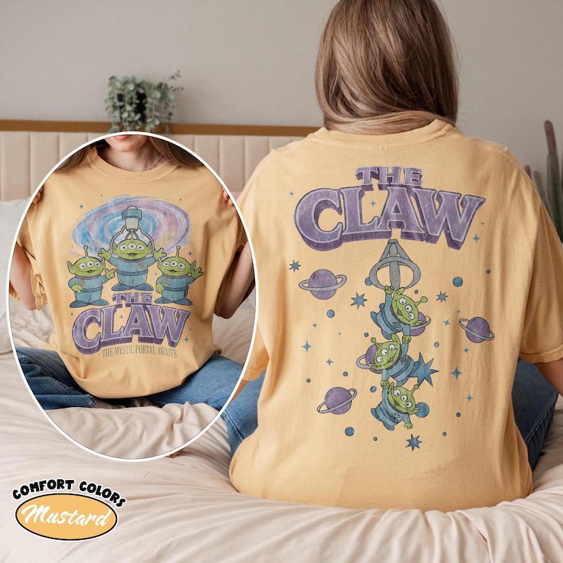 May include: A mustard yellow t-shirt featuring a graphic of three green aliens and a claw machine. The back displays the text THE CLAW with planets and stars. The front graphic includes the text THE CLAW and THE MYSTIC PORTAL AWAITS.