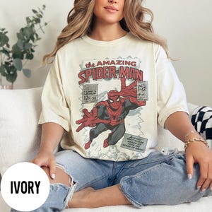 May include: Ivory-colored t-shirt featuring a comic book cover design of Spider-Man. The graphic includes the text "The Amazing Spider-Man," "Marvel Comics Group," and issue details. The superhero is depicted in his classic red and black suit.