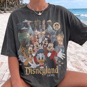 May include: A charcoal gray short-sleeve t-shirt featuring a graphic print of Mickey Mouse, Minnie Mouse, Donald Duck, and Pluto in front of a castle. The text Disneyland Est. 1955 is printed at the bottom of the graphic in a gold-toned font.
