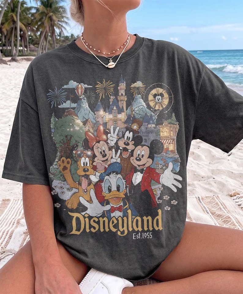 May include: A charcoal gray short-sleeve t-shirt featuring a graphic print of Mickey Mouse, Minnie Mouse, Donald Duck, and Pluto in front of a castle. The text Disneyland Est. 1955 is printed at the bottom of the graphic in a gold-toned font.