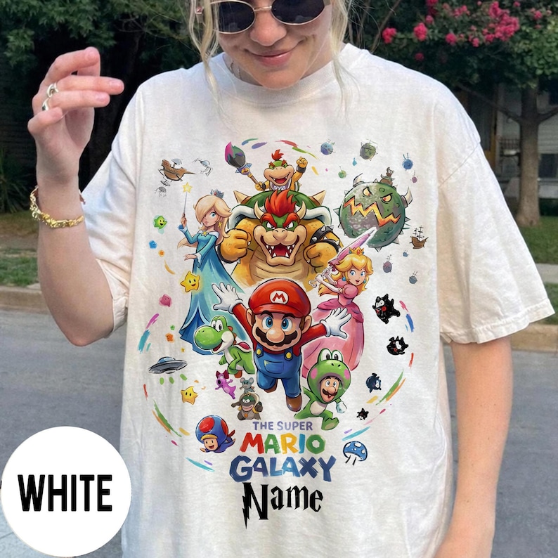 Puede incluir: Camiseta blanca con un dise&ntilde;o colorido de Mario y otros personajes del juego Super Mario Galaxy. El dise&ntilde;o incluye el texto "THE SUPER MARIO GALAXY Name".