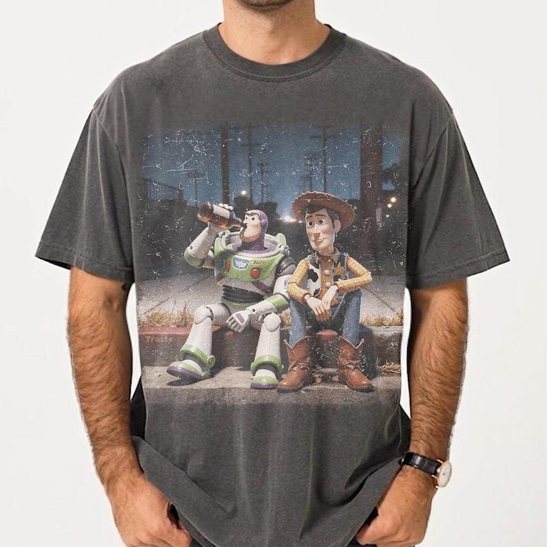 May include: A charcoal grey short-sleeve t-shirt featuring a graphic print of two toy characters sitting on a curb at night. One character is a white and green space ranger holding a brown bottle, and the other is a cowboy wearing a hat and boots.