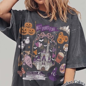 Pode incluir: Uma t-shirt de manga curta cinzento-carvão com uma impressão gráfica com o texto "MOST SPOOKIEST PLACE". O design inclui um castelo, abóboras, um monotrilho, um chapéu de feiticeiro, um balde de pipocas e vários ícones em roxo, laranja e branco.