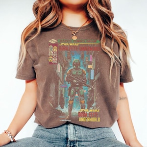 Puede incluir: Una camiseta marr&oacute;n con un gr&aacute;fico de Star Wars. El dise&ntilde;o incluye el texto "Marvel Comics Group Star Wars Boba Fett Underworld" y una ilustraci&oacute;n de Boba Fett en una ciudad. La camiseta es de cuello redondo y manga corta.