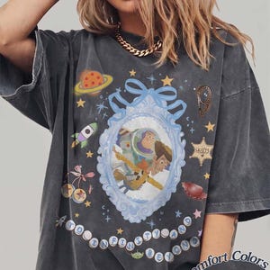 Peut inclure: Un t-shirt gris anthracite oversize avec un cadre bleu clair orn&eacute; au centre contenant Buzz l'&Eacute;clair et Woody. Le motif inclut une plan&egrave;te, une fus&eacute;e, une &eacute;toile de sh&eacute;rif avec le texte "Howdy" et l'inscription "To Infinity and Beyond" en cercles.