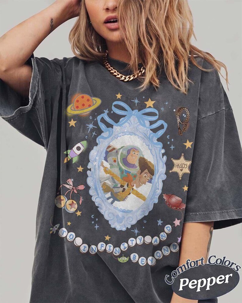 Peut inclure: Un t-shirt gris anthracite oversize avec un cadre bleu clair orn&eacute; au centre contenant Buzz l'&Eacute;clair et Woody. Le motif inclut une plan&egrave;te, une fus&eacute;e, une &eacute;toile de sh&eacute;rif avec le texte "Howdy" et l'inscription "To Infinity and Beyond" en cercles.