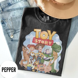 K&ouml;nnte beinhalten: Schwarzes T-Shirt mit der Aufschrift "TOY STORY" und einer Grafik von Figuren aus dem Animationsfilm. Das T-Shirt liegt auf einer blauen Jeans, einer Halskette und einem beigen Hut.