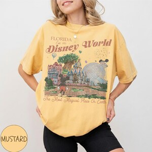May include: Mustard yellow t-shirt featuring a vintage-style graphic of Disney World in Florida. The design includes Mickey Mouse, the castle, and the text "Florida Disney World" and "The Most Magical Place On Earth."