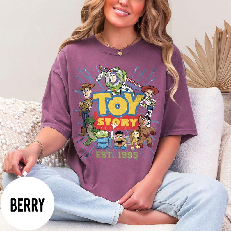 以下が含まれることがあります： ベリー色のTシャツ。アニメ映画「トイ・ストーリー」のキャラクターのグラフィックが特徴です。デザインにはウッディ、バズ・ライトイヤー、ジェシーなどのキャラクターと「Toy Story」と「EST. 1995」の文字が含まれています。