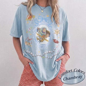 Peut inclure: Un t-shirt bleu clair avec un motif central repr&eacute;sentant Buzz l'&Eacute;clair et Woody dans un cadre ovale orn&eacute;. Le design comprend une fus&eacute;e, une plan&egrave;te, des &eacute;toiles et le texte TO INFINITY AND BEYOND &eacute;crit en cercle autour des personnages.